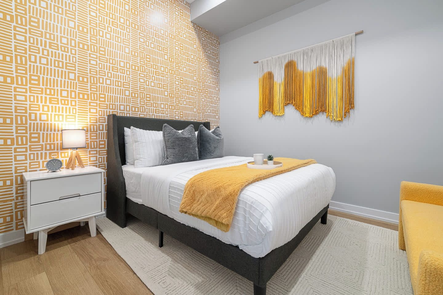 A modern bedroom with a patterned wallpaper, a bed with a white duvet, a bedside table with a lamp, and a yellow throw blanket.