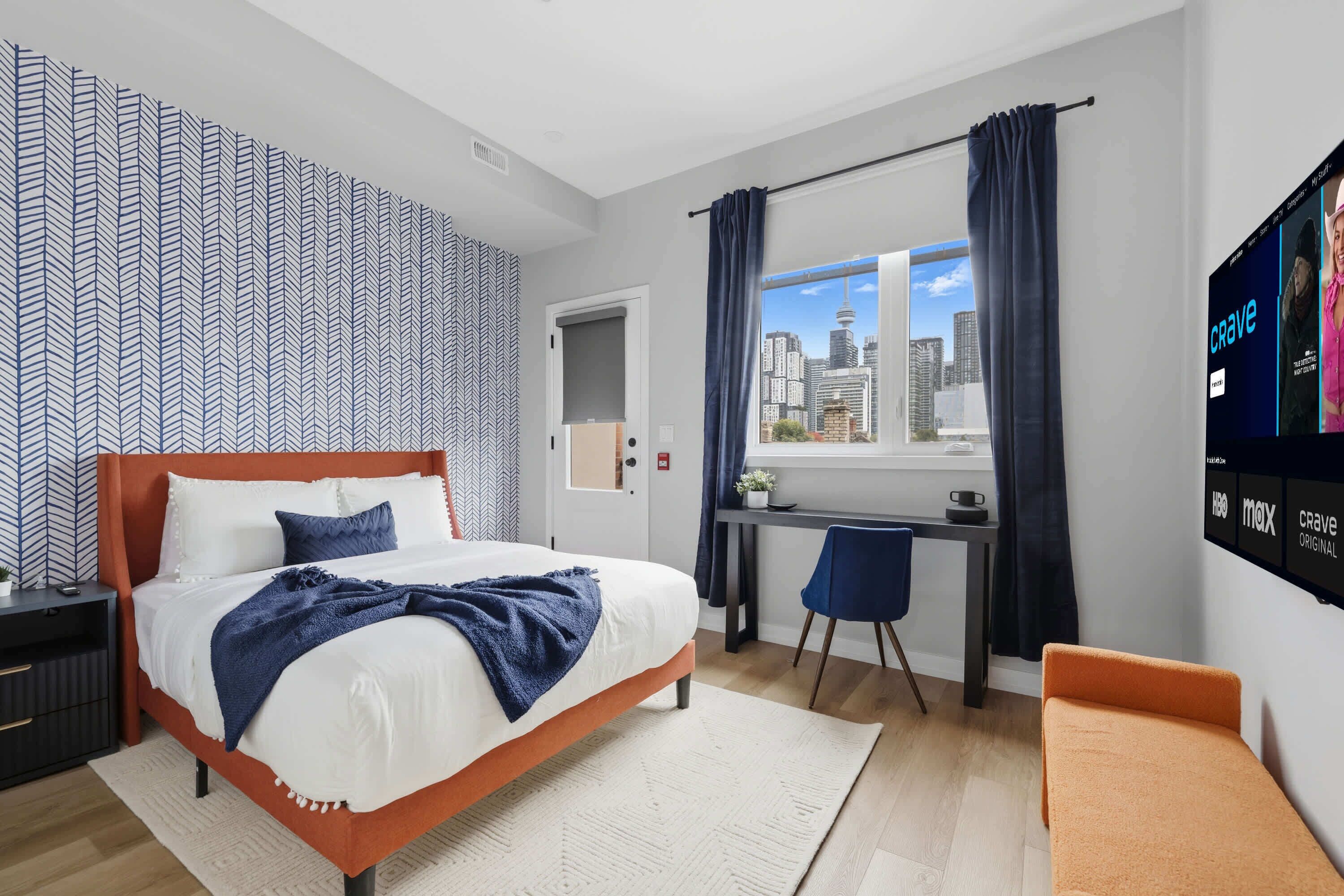 A modern hotel room with a bed, desk, chair, and a TV displaying channels.