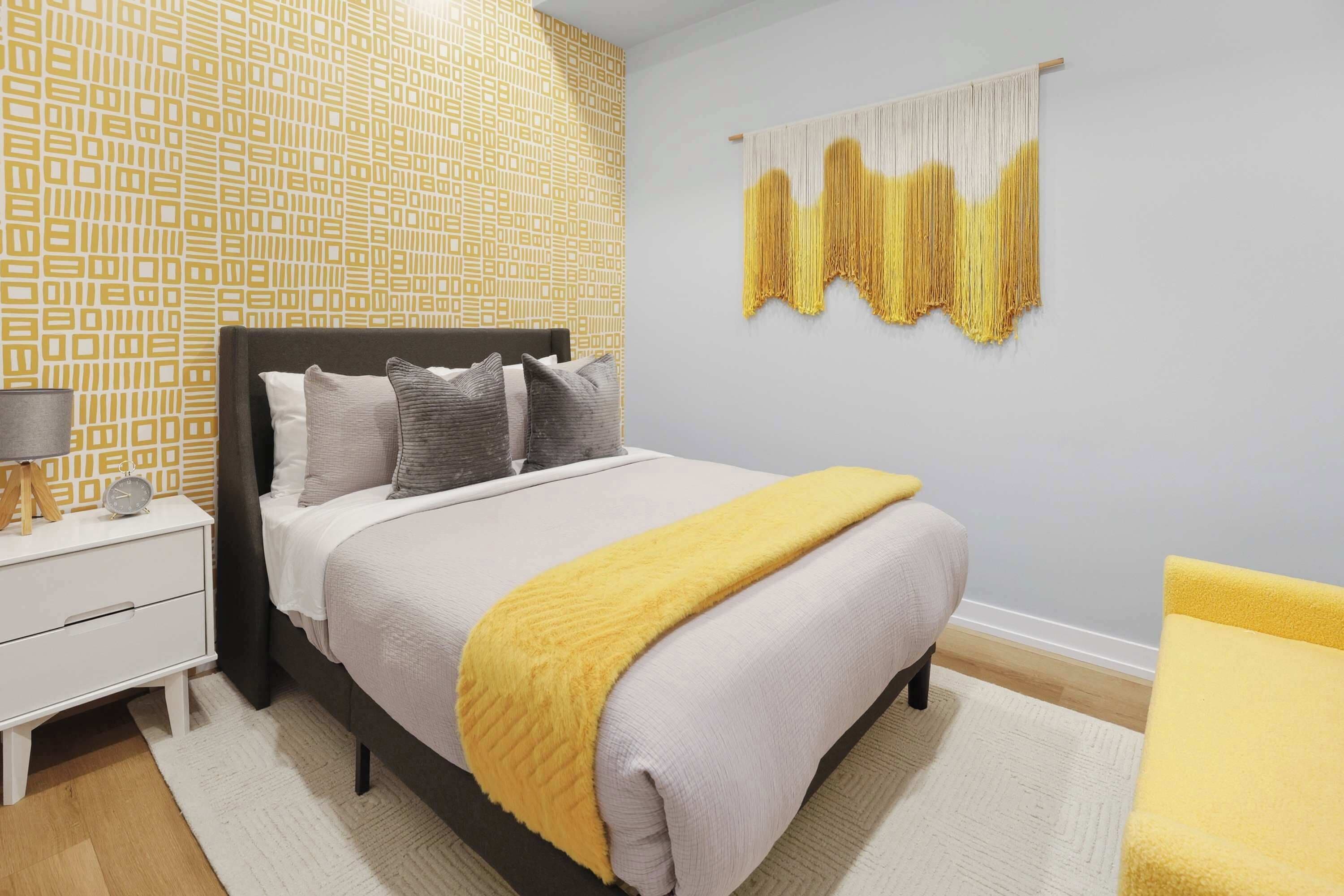 A bedroom with a bed, bedside table, lamp, and a wall hanging.