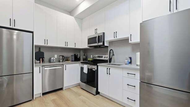 A modern kitchen with stainless steel appliances, white cabinetry, and a wooden floor. - Rare 3BR Gem, Mini Putt Terrace & CN Tower Views! (Toronto)