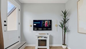 A modern living room with a mounted television displaying the 'Crave' channel, a white console table with decorative items, a tall green plant, and a framed picture on the wall. - Rare 3BR Gem, Mini Putt Terrace & CN Tower Views! (Toronto)