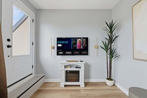 Ein modernes Wohnzimmer mit einem an der Wand befestigten Fernseher, der den Sender 'Crave' zeigt, einem weißen Konsolentisch mit Dekoration, einer hohen, grünen Pflanze und einem gerahmten Bild.