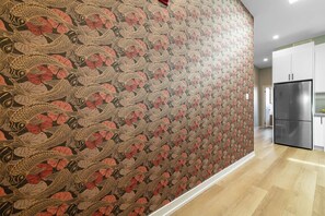 A hallway with patterned wallpaper, a kitchen area with white cabinets, and a stainless steel refrigerator.