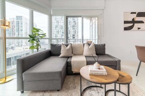 A modern living room with a grey sectional sofa, a small round table, and a large window offering a city view.