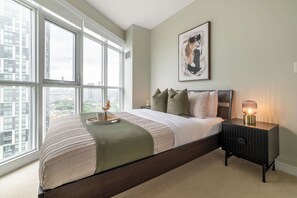 Ein Schlafzimmer mit großem Fenster, einem Bett mit Kissen, einem Nachttisch mit Lampe und einem gerahmten Bild an der Wand.