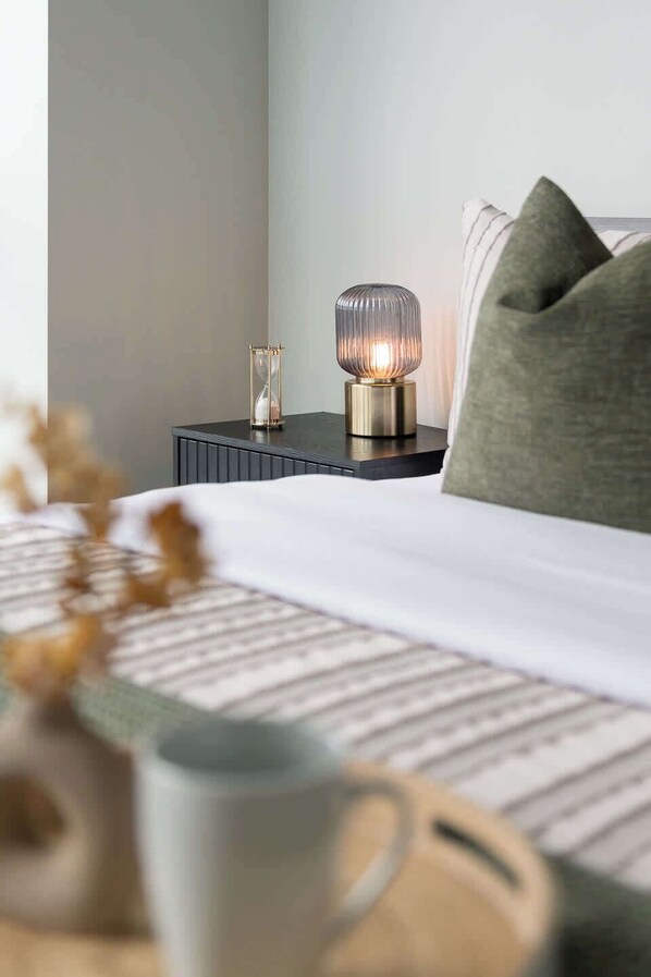 A neatly made bed with a white and patterned bedspread, a bedside table with a lamp, and a decorative vase with dried flowers.