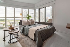 A modern bedroom with a large bed, a small table, and a view of a cityscape through large windows. - CityPlace 2BR Escape | Balcony & 2 Bathrooms (Toronto)