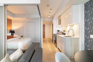 A modern hotel room with a bed, bedside table, lamp, and a view into the adjacent kitchen area. - Modern 1BR, Designer Condo, Sleeps 4 | Parking Available (Toronto)