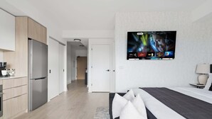 A modern hotel room with a large flat-screen TV mounted on the wall, a bed with a grey and white duvet, and a kitchenette with a stainless steel refrigerator.