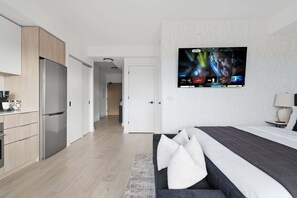 Une chambre d’hôtel moderne dotée d’un grand téléviseur à écran plat fixé au mur, d’un lit avec une couette grise et blanche, et d’une kitchenette équipée d’un réfrigérateur en acier inoxydable.