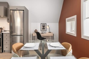 Una cocina moderna con un refrigerador de acero inoxidable, una mesa de comedor con dos sillas, y un escritorio pequeño con una silla y una lámpara.