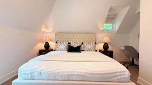 Ein Schlafzimmer mit einem großen Bett, zwei Nachttischen mit Lampen, einem grauen Bürostuhl und einer schrägen Decke mit Dachfenster.