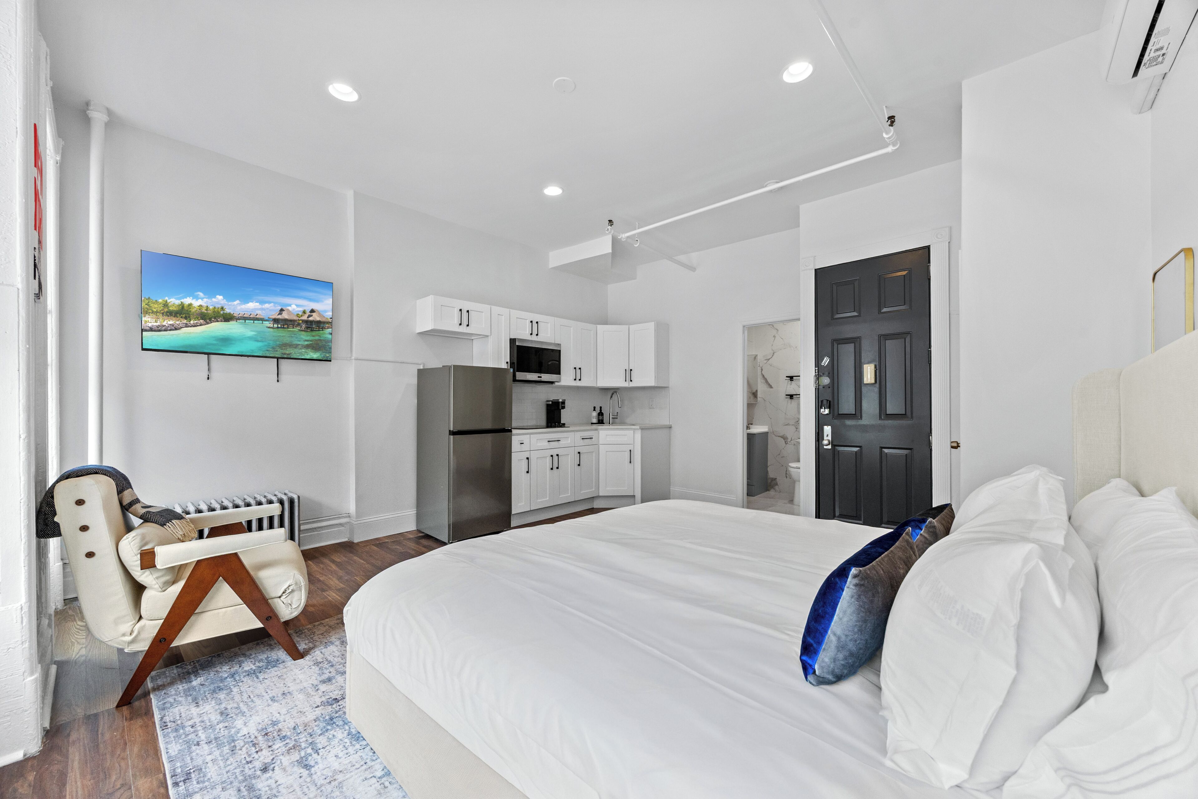 A modern bedroom with a large bed, a flat-screen TV, a kitchen area, and a bathroom.