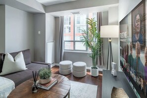 Ein modernes Wohnzimmer mit einer Couch, Hocker, einem Couchtisch, einer Lampe und einem großen Fenster mit Vorhängen.