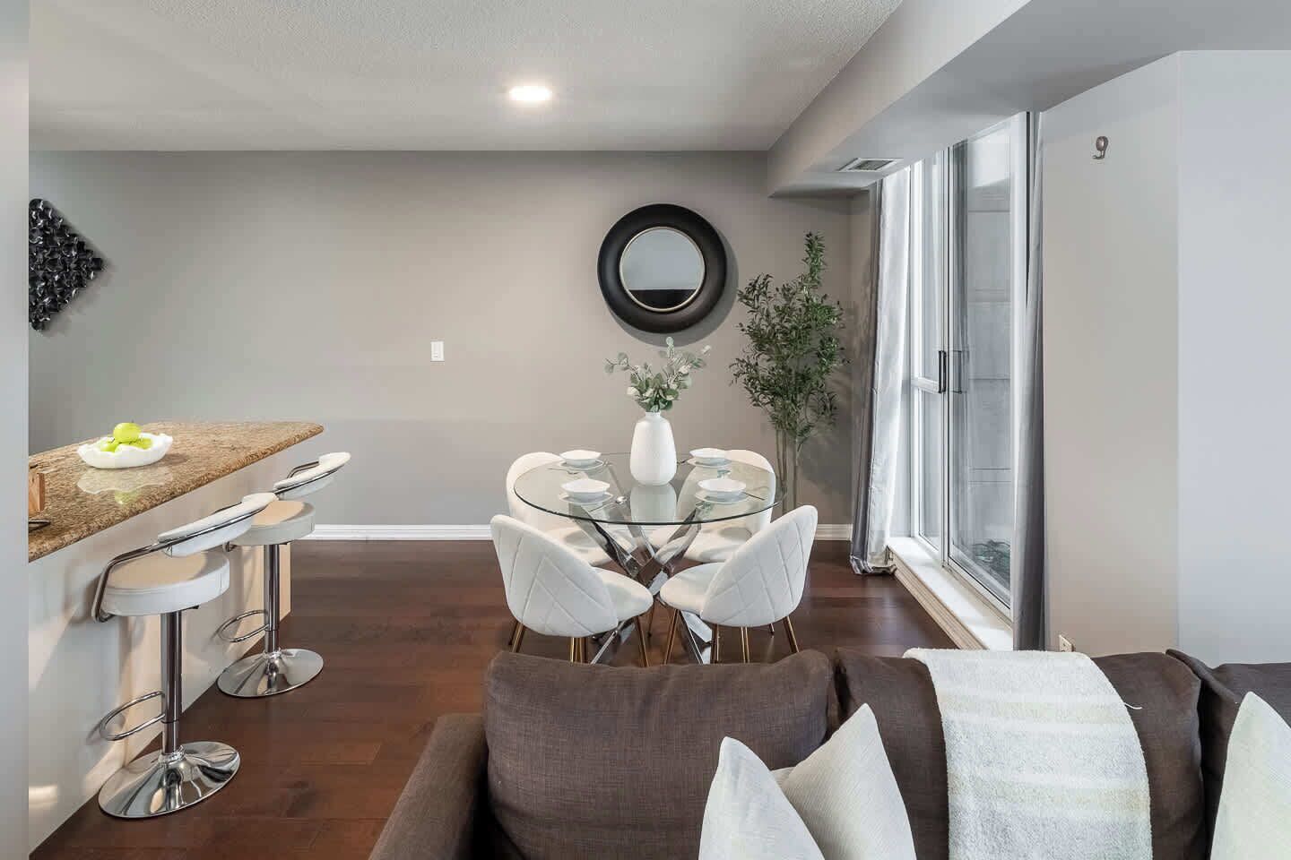 A modern living room with a glass dining table, white chairs, a brown sofa, and a large window.
