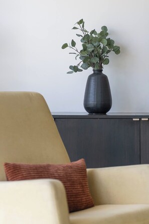 Ein Zimmer mit einem beige gefärbten Sessel, einer dunkelbraunen Vase mit Grünpflanzen und einer dunklen Holzschrankwand an einer weißen Wand.