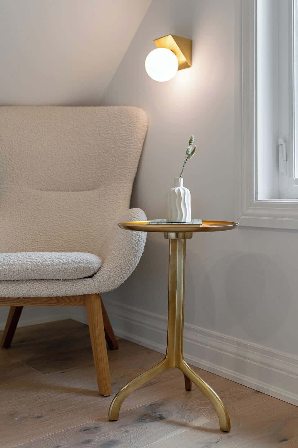 A room with a beige armchair, a small round table with a white vase, and a wall-mounted light fixture.