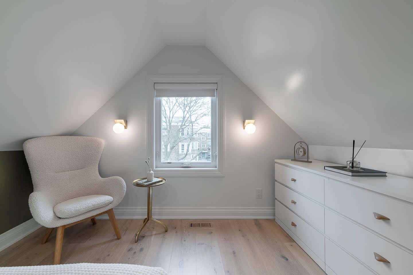 A room with a sloped ceiling, a window with blinds, a white chair, a small round table with a vase, and a white dresser with drawers.