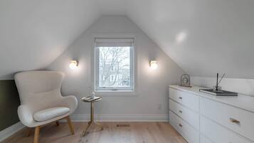 A room with a sloped ceiling, a window with blinds, a white chair, a small round table with a vase, and a white dresser with drawers.