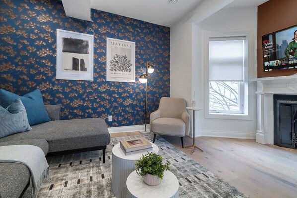 A living room with a sofa, armchair, and a fireplace. There are wall art pieces and a television mounted on the wall.