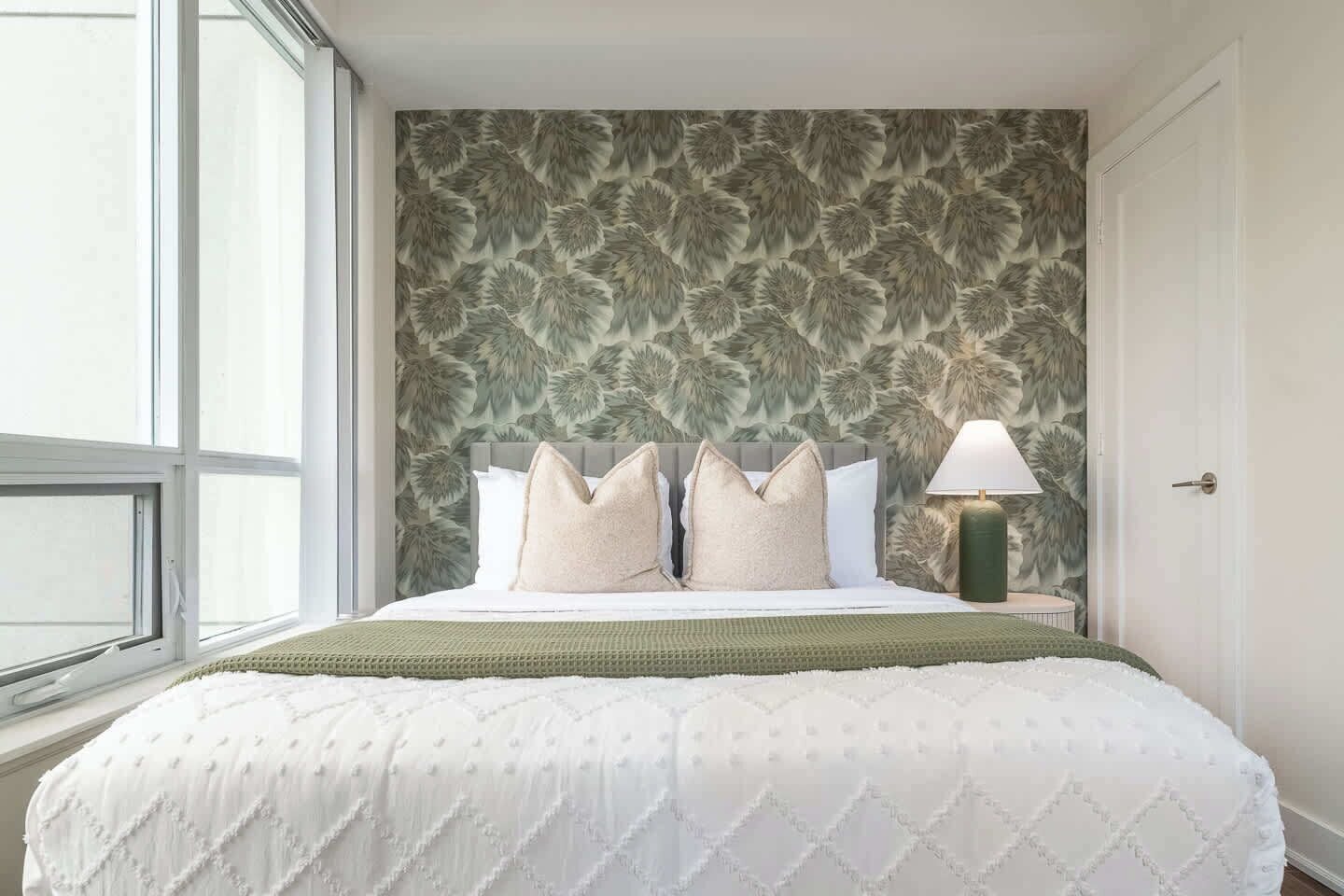 A bedroom with a large bed, a window, a patterned wallpaper, and a nightstand with a lamp.