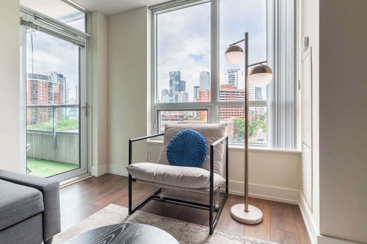 A modern apartment with a balcony, a chair with a cushion, a floor lamp, and a view of the cityscape.