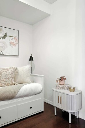 A bedroom with a white bed, a bedside lamp, a small white cabinet with a crown decoration, and a floral painting on the wall. - Downtown Financial District Gem Sleeps 5 55"TV (Toronto)
