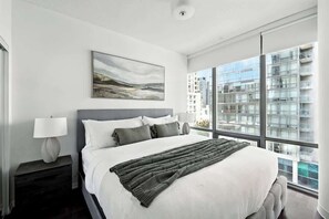 A modern hotel room with a large bed, a nightstand with a lamp, a painting on the wall, and a view of a cityscape through large windows. - Downtown Financial District Gem Sleeps 5 55"TV (Toronto)