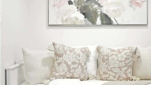A white sofa with floral-patterned pillows and a plain wall with a framed floral painting. - Downtown Financial District Gem Sleeps 5 55"TV (Toronto)
