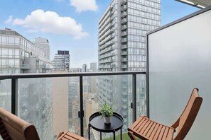 Un balcón con vistas a edificios altos, una pequeña mesa con una planta y dos sillas de madera.