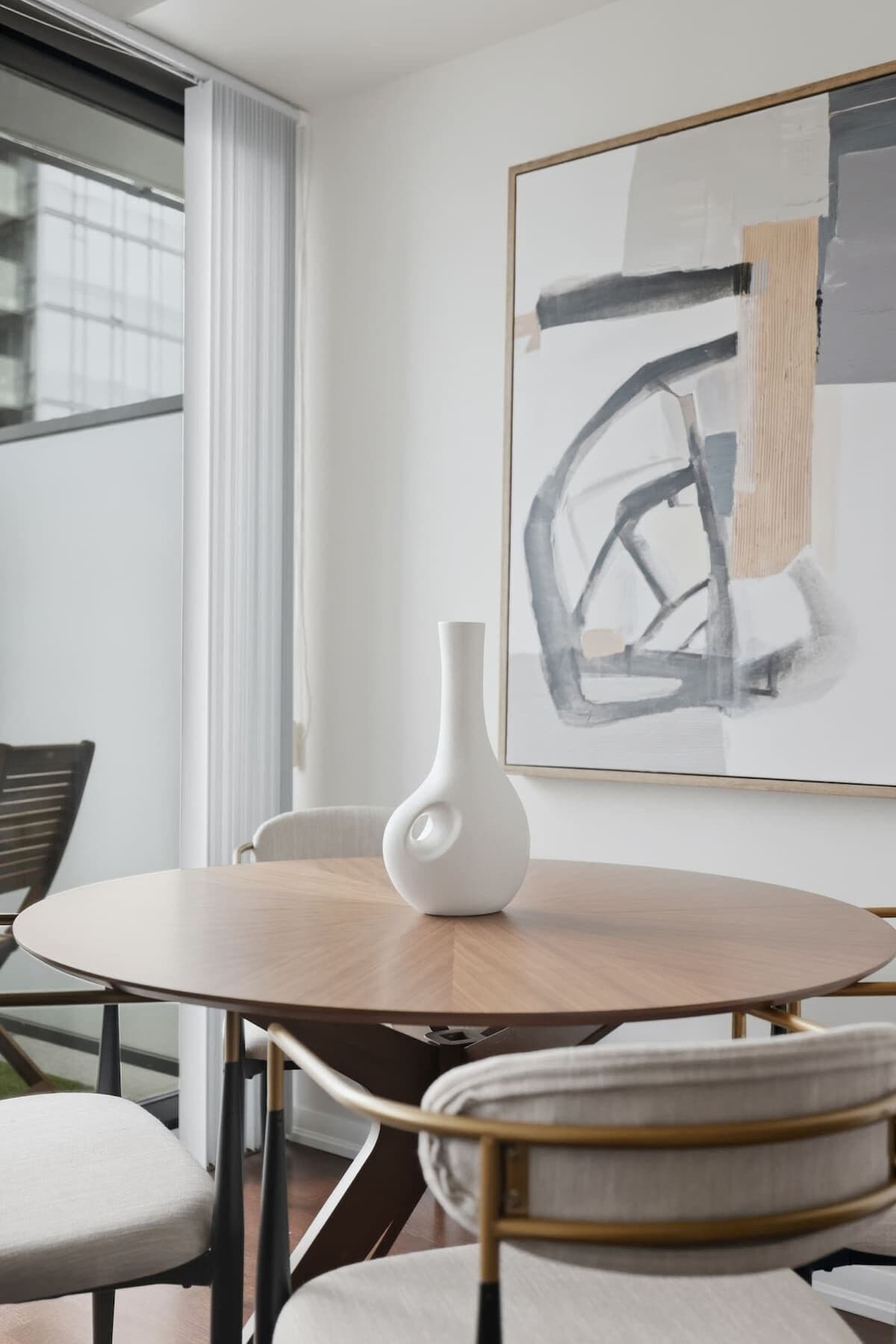Een ronde eettafel met een witte vaas, een lijst met abstract schilderij en een raam met jaloezieën.