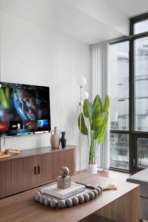 Ein modernes Wohnzimmer mit einem an der Wand befestigten Flachbildfernseher, einem Holztisch mit Dekoration, einer Topfpflanze und Blick auf die Stadt durch große Fenster.