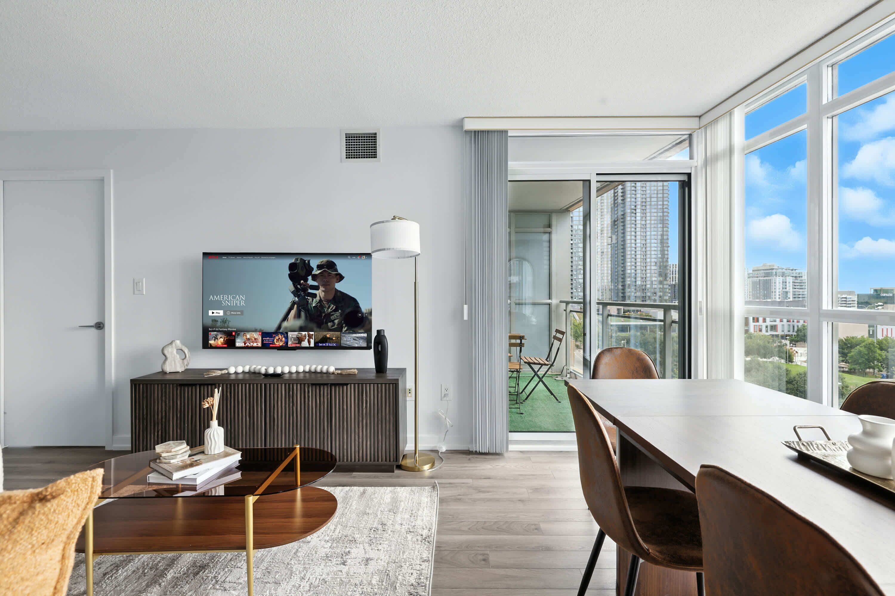 A modern living room with a flat-screen TV, a dining area with a table and chairs, and a large window offering a city view.