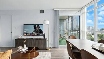 Uma sala de estar moderna com televisão de tela plana, uma área de jantar com mesa e cadeiras, e uma janela grande com vista para a cidade.