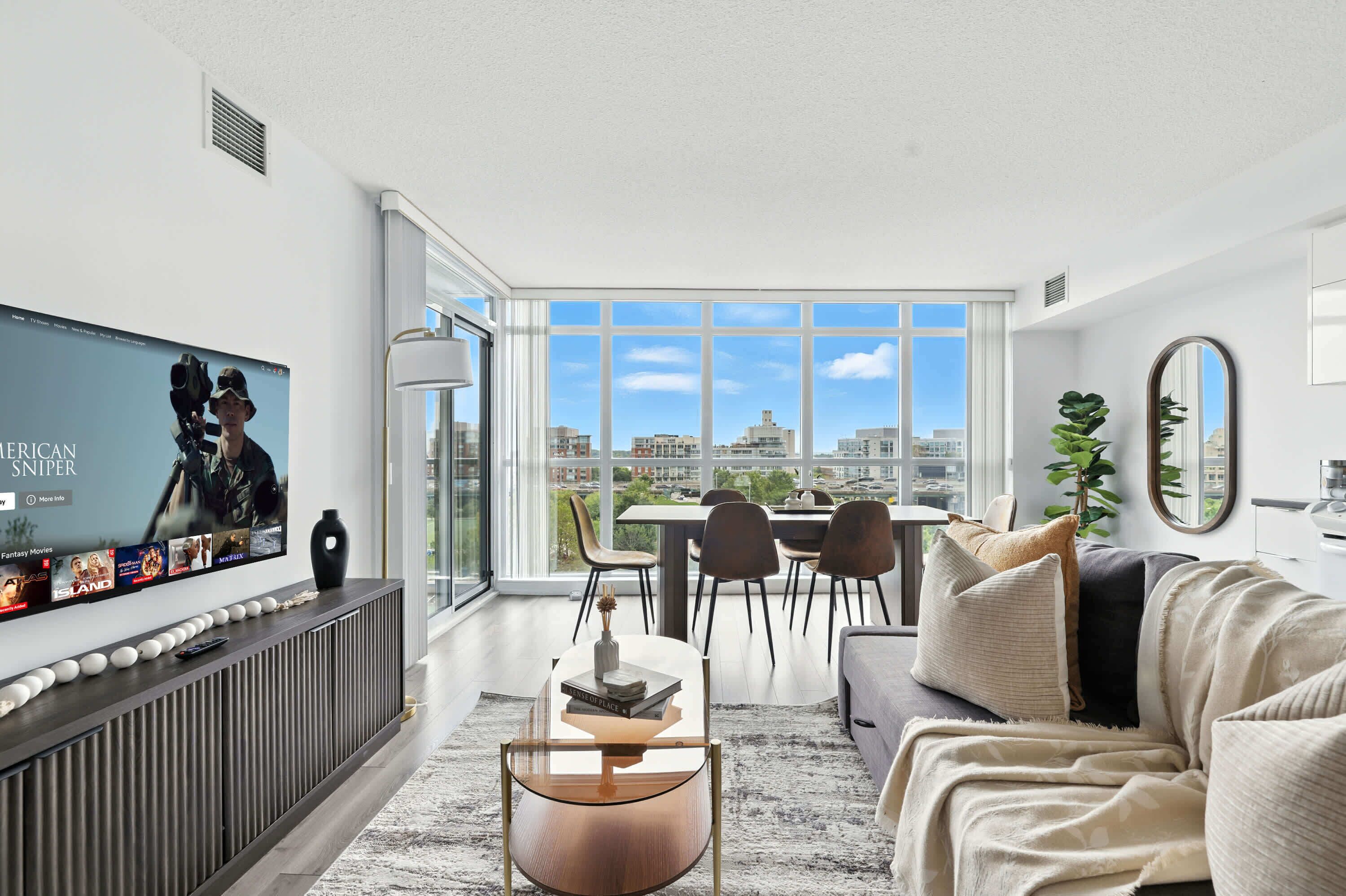 A modern living room with a large TV, a dining area, and a city view.
