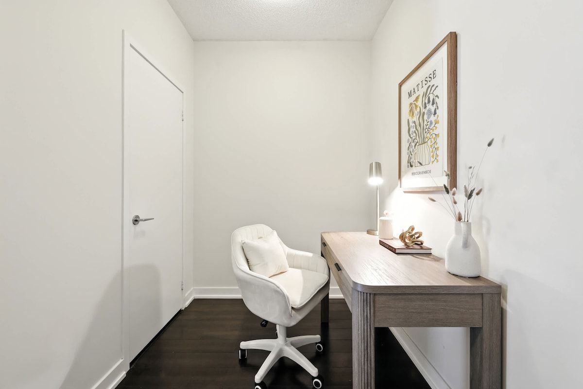 A small, clean room with a white door, a white office chair, a wooden desk, and a framed artwork on the wall.