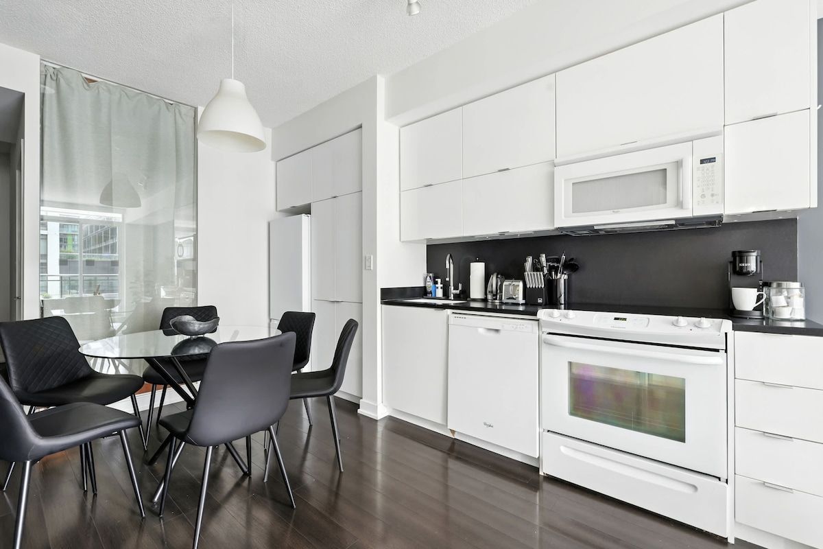 Une cuisine moderne avec des meubles blancs, un plan de travail noir et un coin repas comprenant une table en verre et des chaises noires.