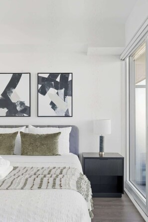 A modern bedroom with a large bed, two abstract paintings, a bedside lamp, and a view through a large window. - Urban 2BR Suite near Yonge-Dundas | Private Balcony | Parking Available (Toronto)