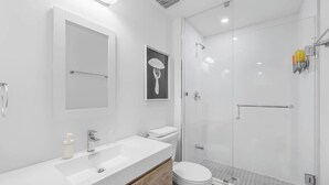 Un baño moderno con lavamanos blanco, tocador de madera, inodoro y zona de ducha con mampara de vidrio.