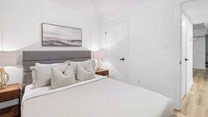 Un dormitorio con cama, mesitas de noche, lámpara, puerta y un cuadro colgado en la pared.