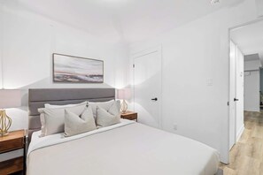 A bedroom with a bed, bedside tables, a nightstand, a lamp, a door, and a wall-mounted picture. - Cozy 2BR Lower-Level Suite | Fast Transit! (Toronto)