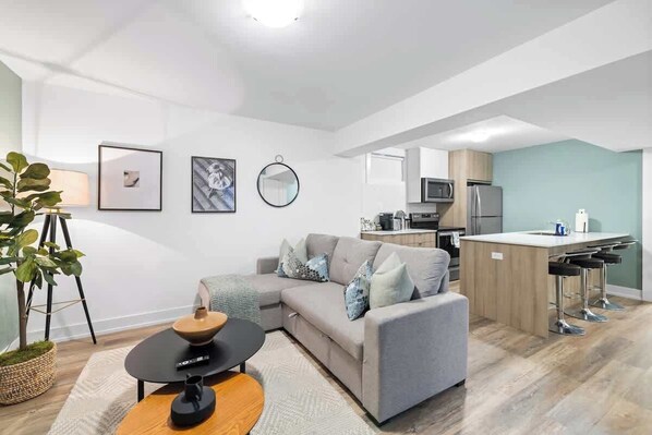 A modern living room with a sofa, a coffee table, and a small kitchen area with a microwave and a refrigerator. - Cozy 2BR Lower-Level Suite | Fast Transit! (Toronto)