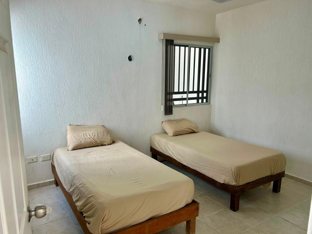 Une chambre avec deux lits simples, une fenêtre avec des stores verticaux et une porte.