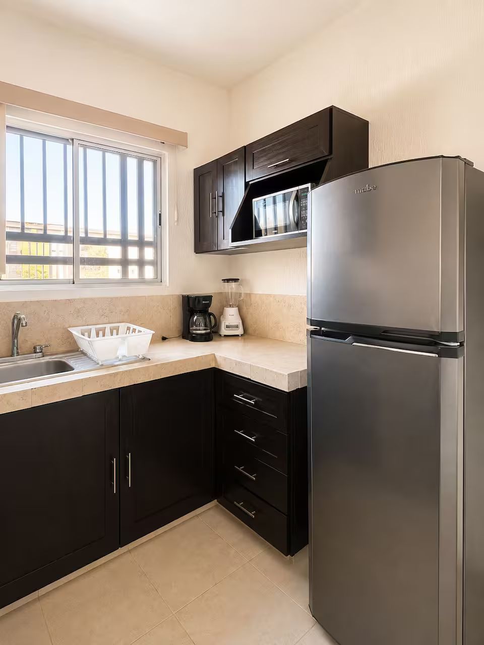 A kitchen with a stainless steel refrigerator, a coffee maker, and a window with blinds.