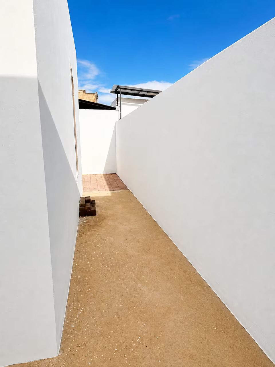 A narrow corridor with white walls and a brown floor, leading to a building with a visible roof.