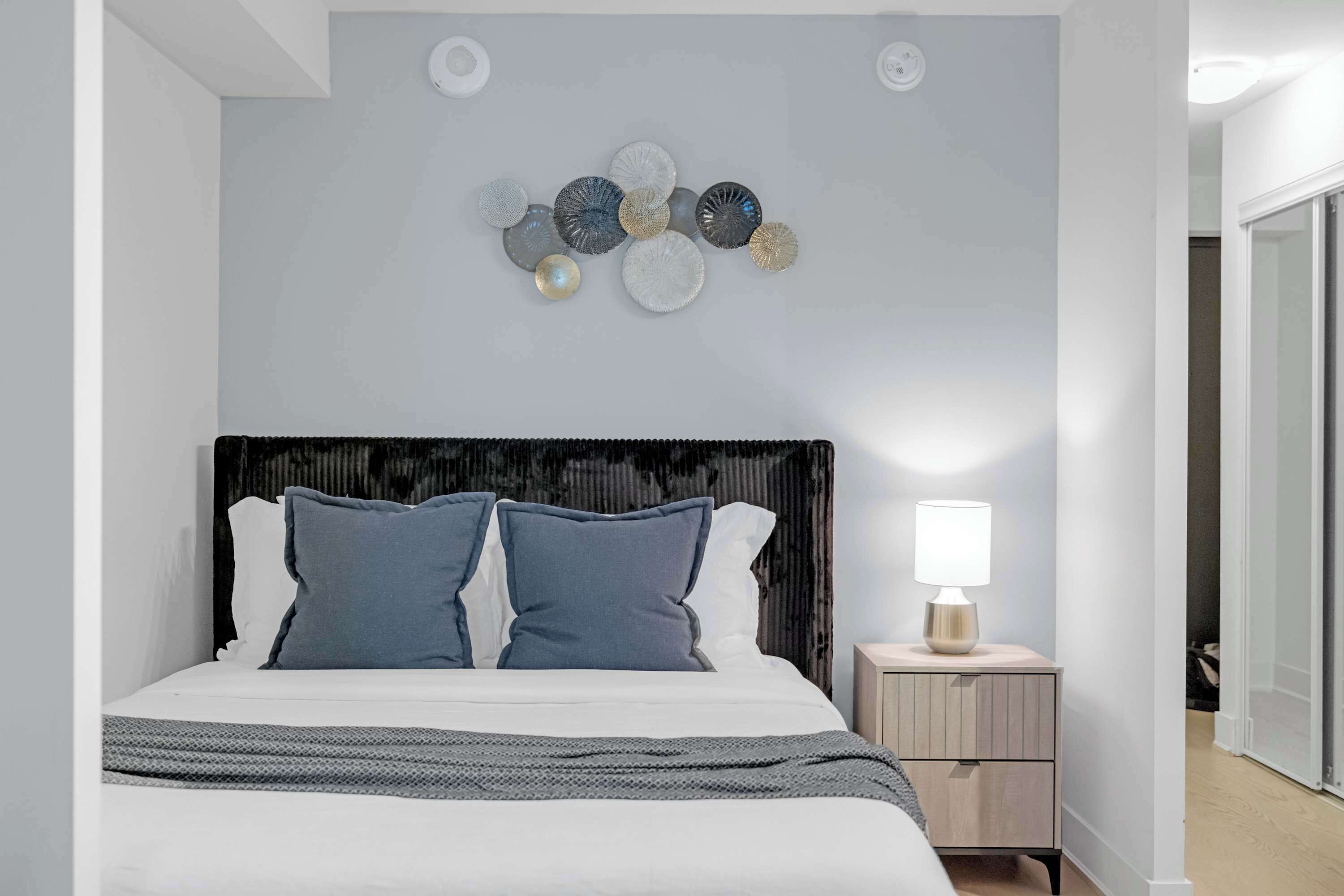 A modern bedroom with a bed, pillows, a bedside table with a lamp, and a decorative wall piece.