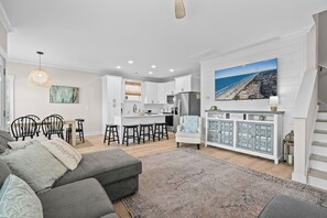 Un salon moderne avec un canapé gris, un coin repas avec des chaises, une cuisine avec des meubles blancs et un grand téléviseur diffusant une scène de plage.