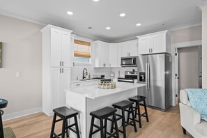 A modern kitchen with white cabinets, a stainless steel refrigerator, and a central island with bar stools. - Driftwood Dreams at Ocean Lakes - w Golf Cart (Myrtle Beach)
