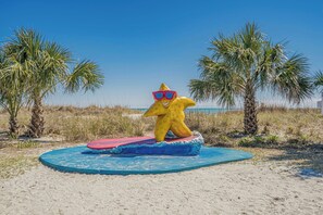 Eine Strandansicht mit einer großen, gelben Seestern-Statue, die eine Sonnenbrille und einen roten Hut trägt und auf einer blauen Matte sitzt.