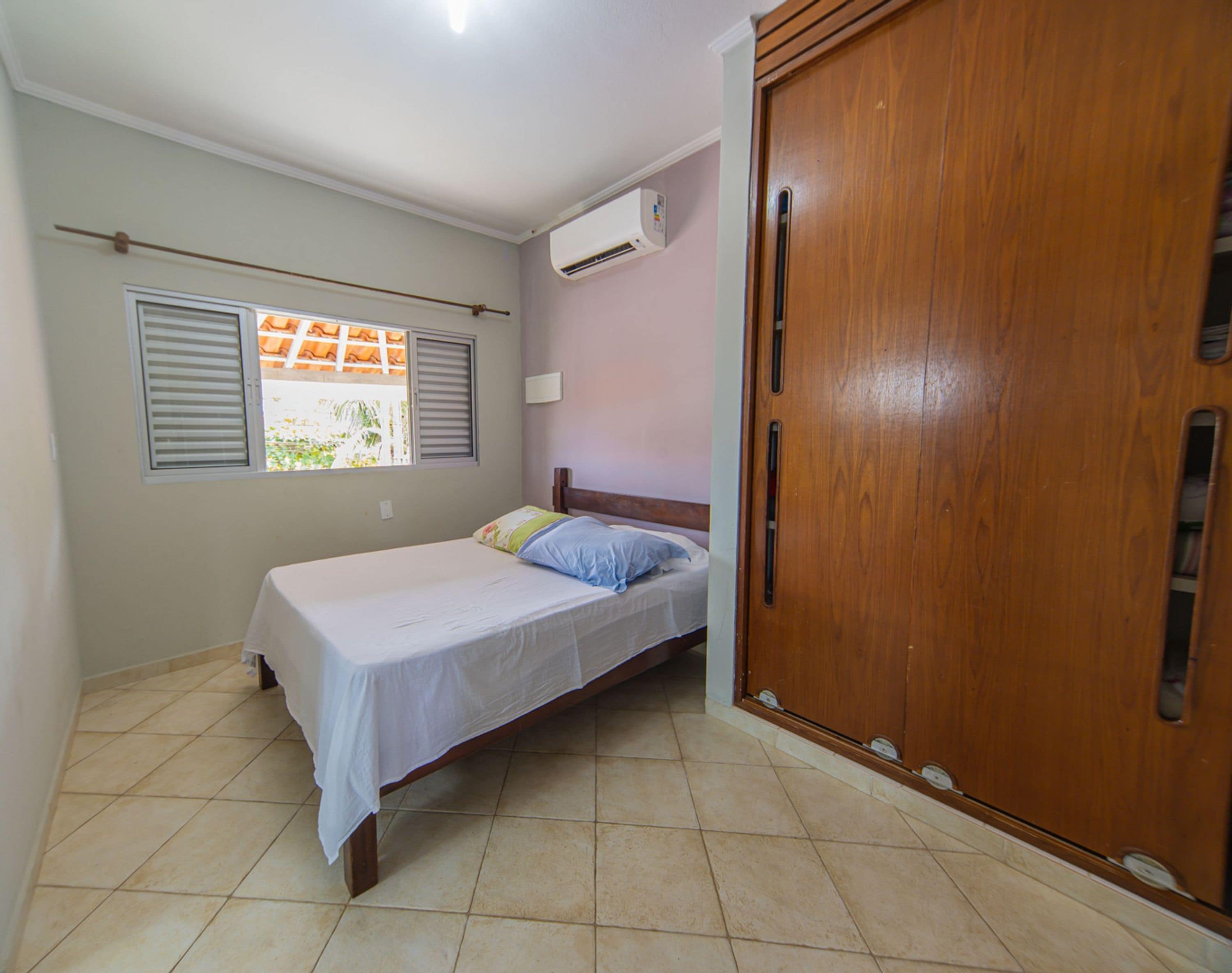 A bedroom with a bed, a wooden wardrobe, an air conditioning unit, and a window with shutters.